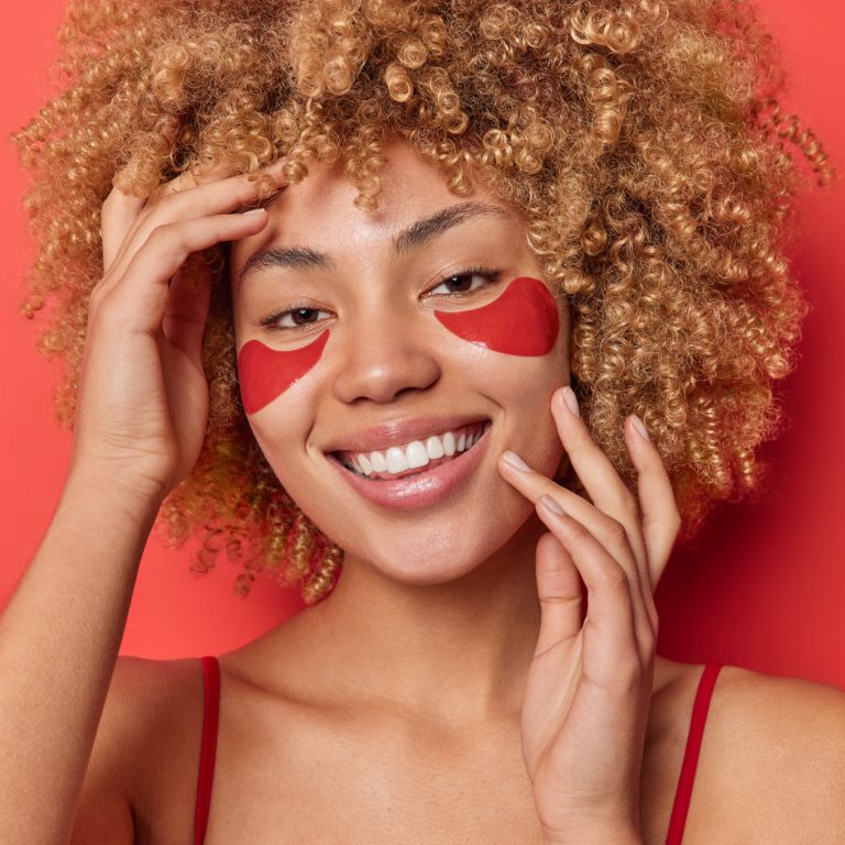Photo of positive woman with curly blonde hair applies collagen patches under eyes for reducing wrinkles and fine lines smiles toothily isolated over red background. Skin care and wellness concept